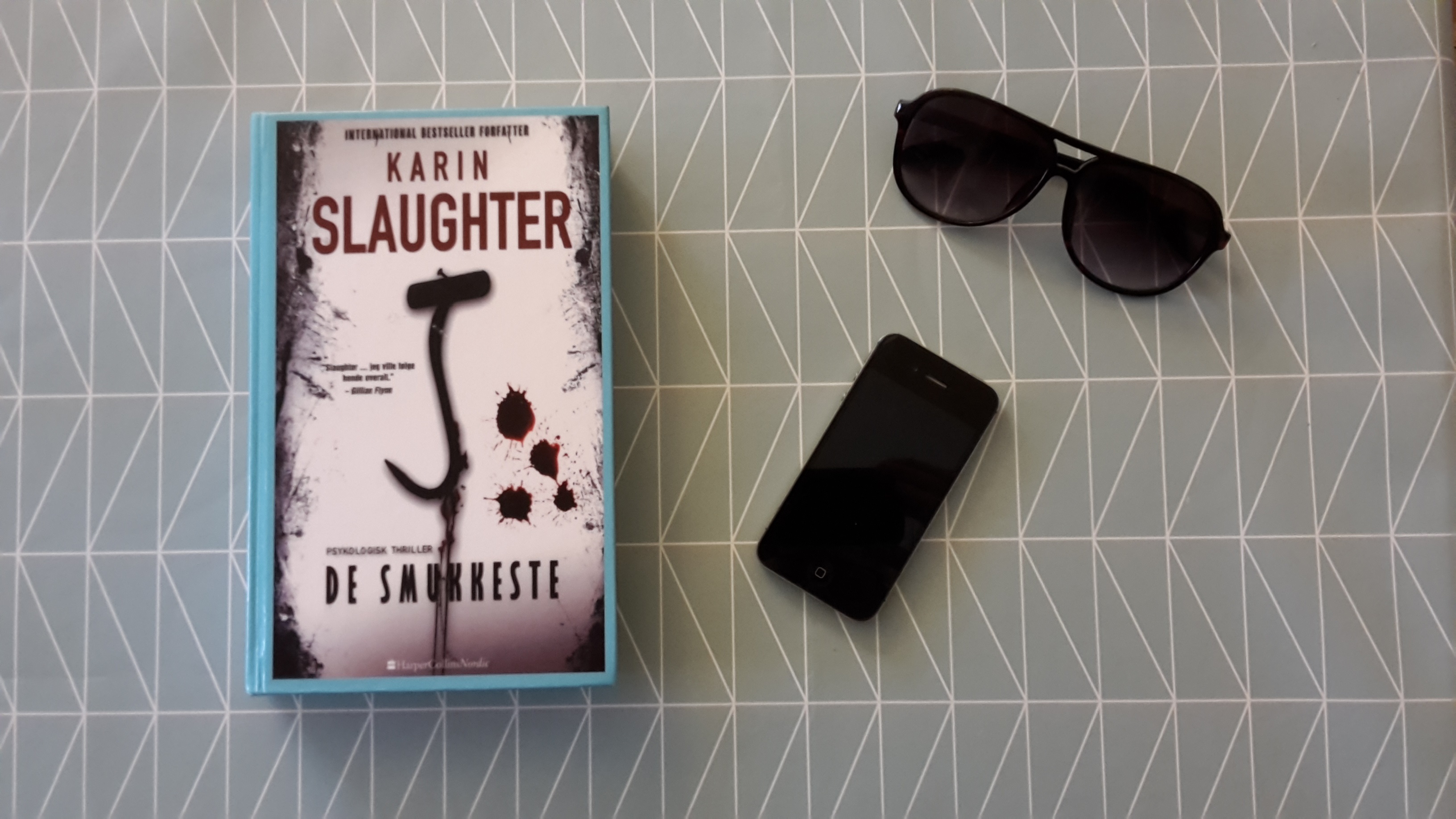 The book Pretty Girls by Karin Slaughter lies on a table with a smart phone and a pair of sunglasses. The table is covered in a blue table cloth.