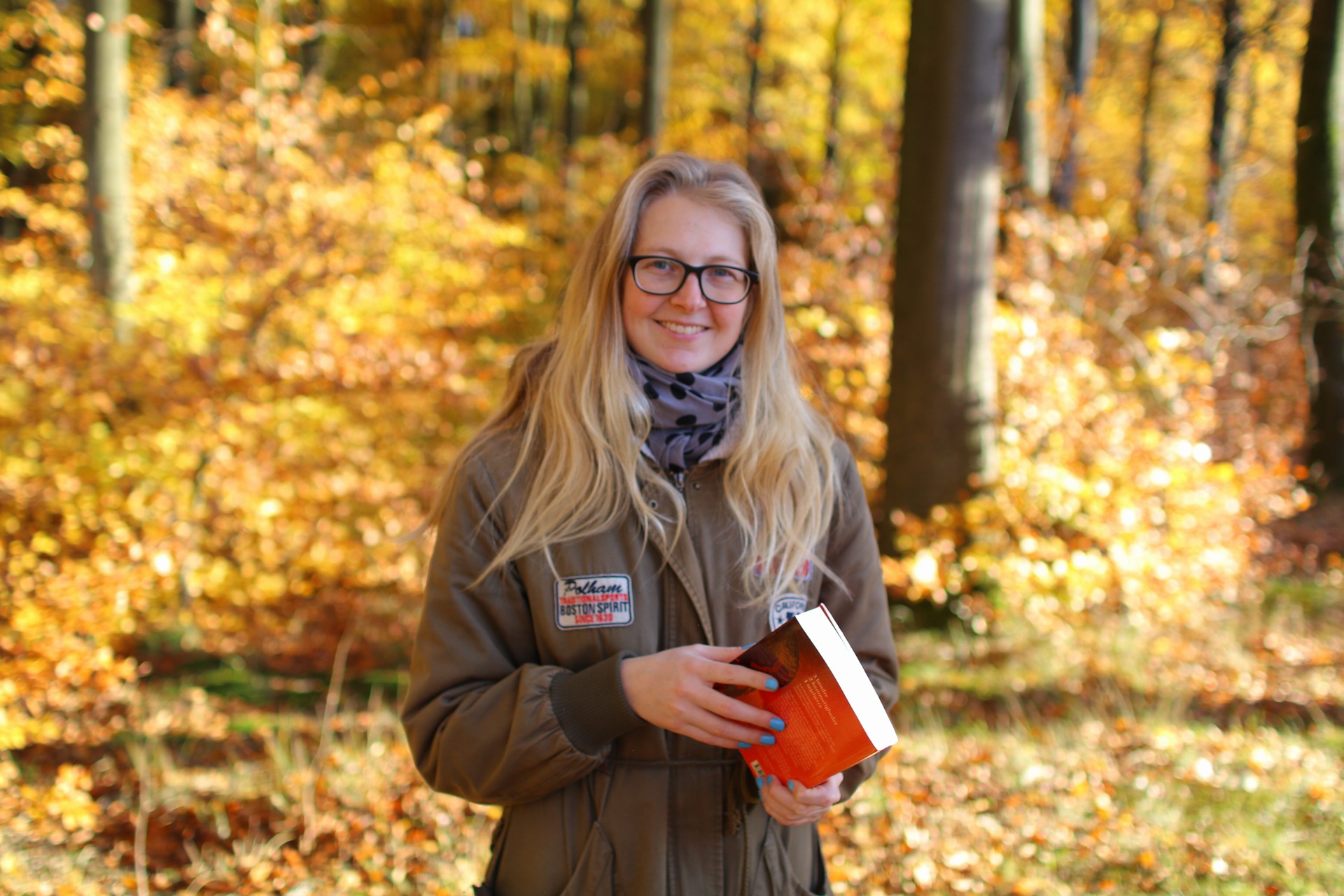 Billedet viser mig, der står i skoven med en orange bog i hånden. I baggrunden ser man træer og blade i efterårets gule og orange farver.